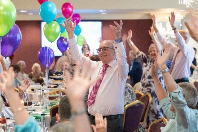 A group of people in a room with balloons for the Volunteer Celebration Event with the focus on one man waving his arms in the arm dressing in a pink shirt with a dark pink tie