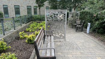 A bench and chairs sat in the middle of a brick walkway with lots of greenery around including trees. To the side of the path is two large stood up metal frames with lots of metal leaf cut outs which is a beautiful pieces of art.