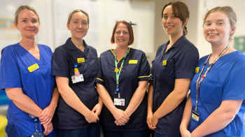 An image of five Chemotherapy members of staff stood smiling for the camera. Two staff members are wearing blue scrubs while two staff members are wearing all Navy blue tunics and the final staff member is wearing a blue tunic with yellow piping.