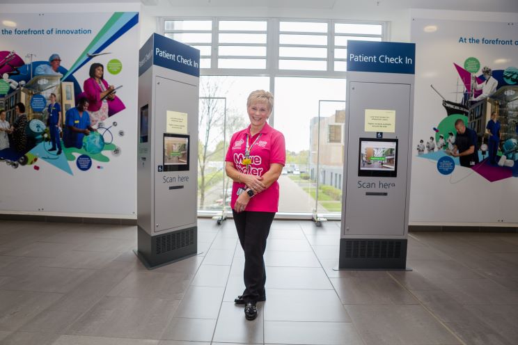 Jill in the Brunel atirum next to the patient check-in kiosks. She is wearing a bright pink Move Maker polo shirt and smiling. 