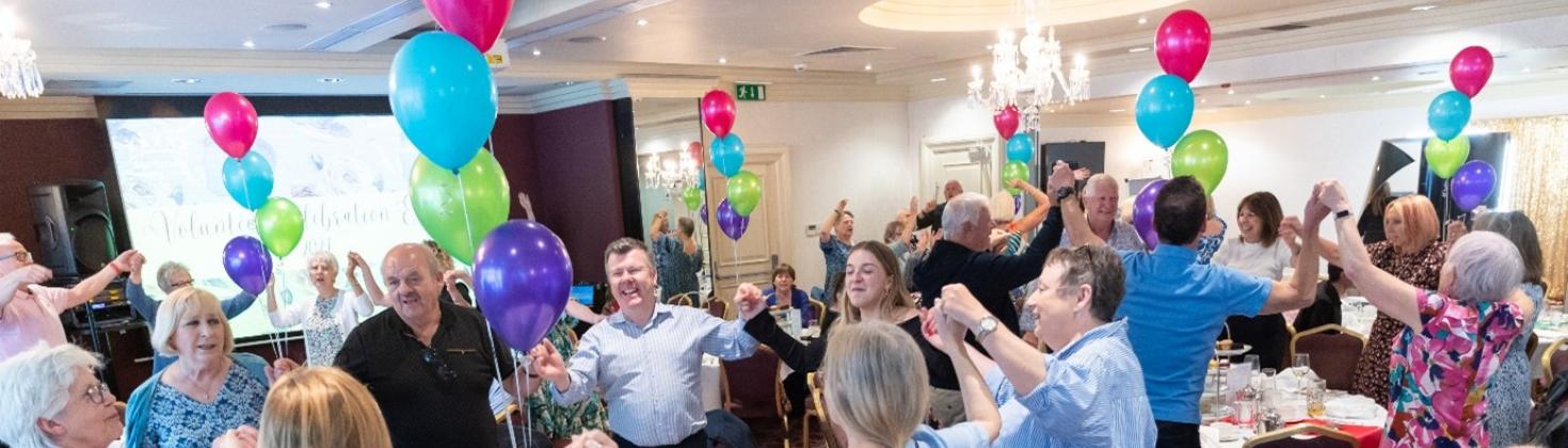 A group of people dancing holding hands during the Volunteer Celebration Event. There are colourful balloons raising up from each of the tables