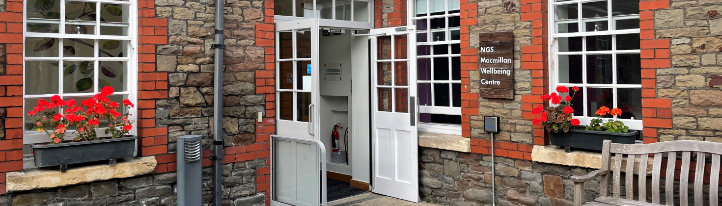 The entrance to the Macmillan Wellbeing Centre. It is a grey stone building with thee red brick window surrounds and white double doors. By the entrance there is a bench.