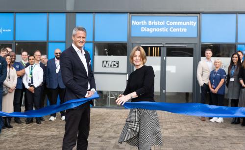 Two people stand in the forefront, cutting a blue ribbon. Behind them is a building with a sign above the door reading "North Bristol Community Diagnostic Centre"