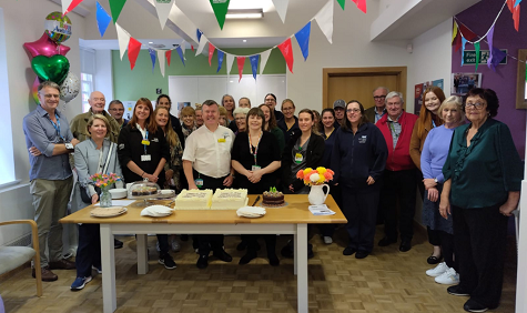 A group of people stand behind a table. There is a large cake on the table. Bunting above and balloons in the background