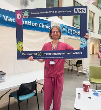 A man in pink scrubs holds up a blue selfie frame with a message encouraging people to get vaccinated.