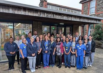 A large group of people, some in scrubs of varying colours, stand, smiling, outside a building with the sign "Bristol Breast Care Centre" above them