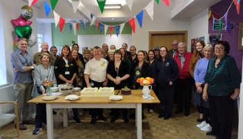 A group of people stand behind a table. There is a large cake on the table. Bunting above and balloons in the background